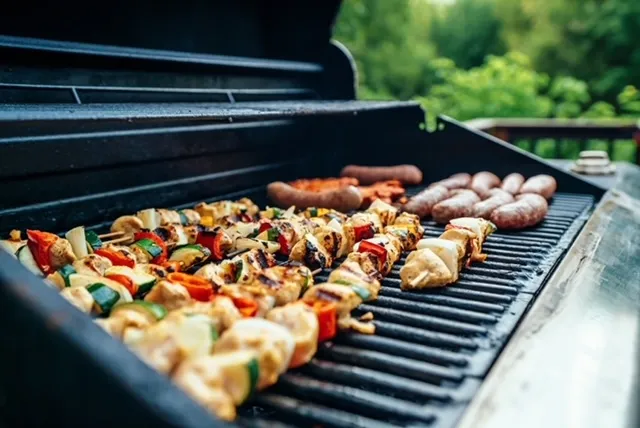 Auf dem Bild ist ein Grill zu sehen, auf dem Bratwürste und verschiedene Fleisch- und Gemüse-Spieße gegrillt werden.